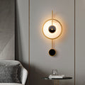Brass ring wall light with a lit alabaster disc and black stone centre, on a grey living room wall.