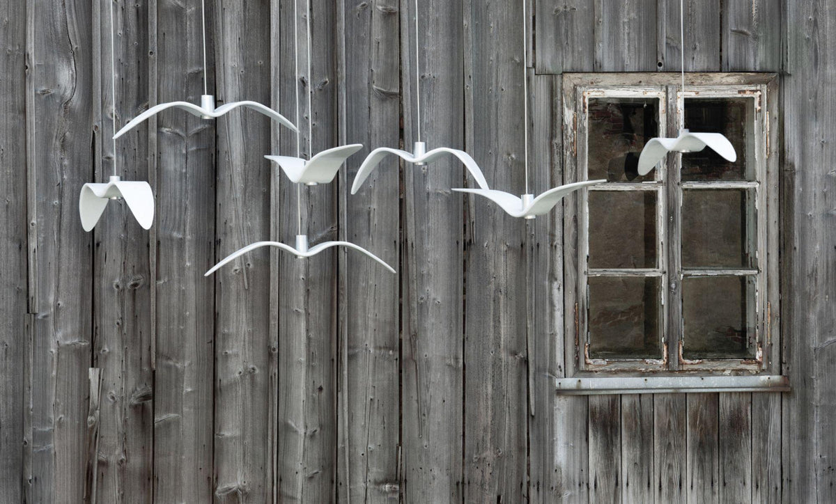 White bird-shaped pendants from this Clérachic fixture hang in a row along a weathered wooden plank wall beside a window.