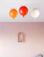 Against a soft pink wall, three Clérachic balloon lights in orange, red and white hang beside a small wooden house shelf.