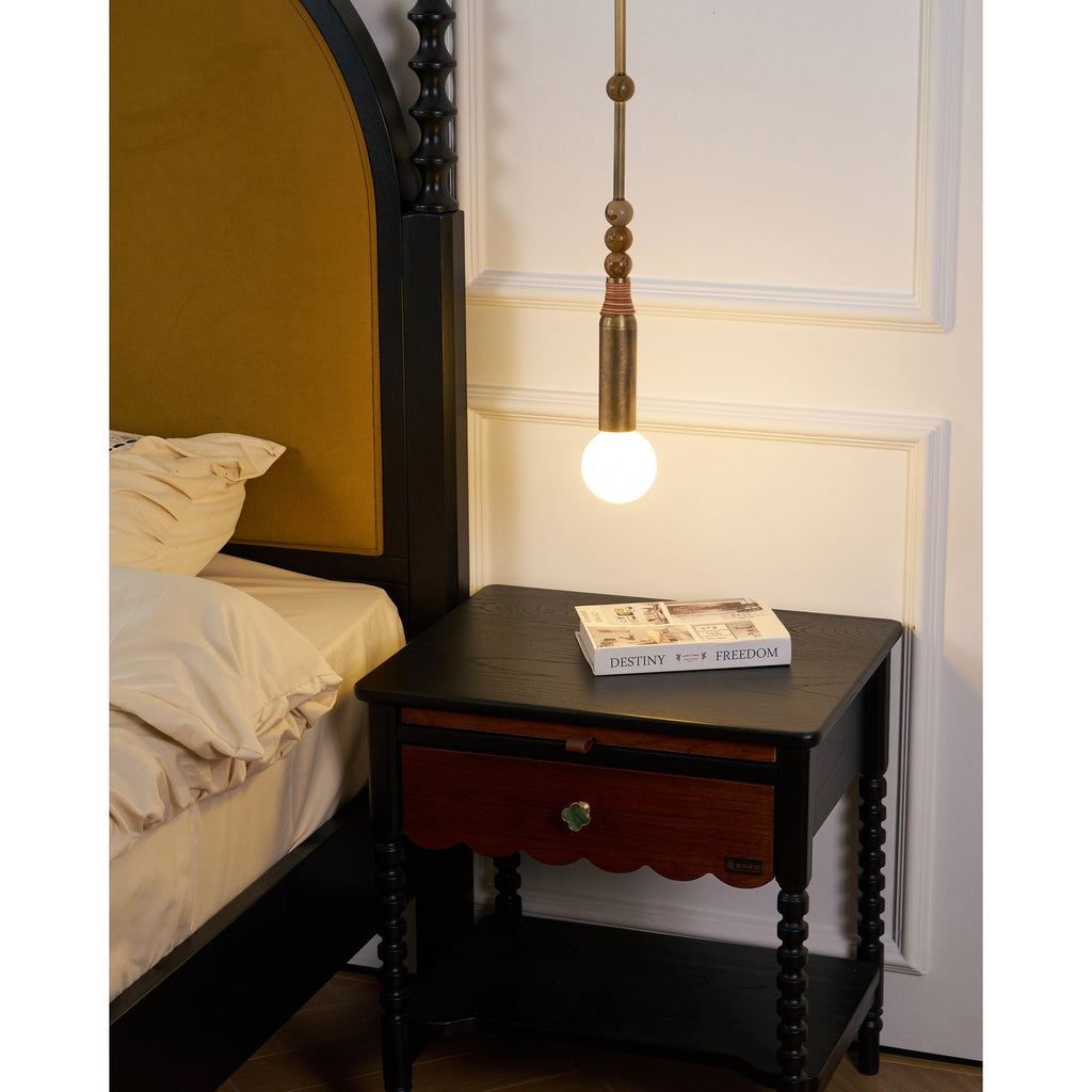 The Clérachic pendant light lit above a dark nightstand, with the bed's arched, mustard-upholstered headboard behind it.