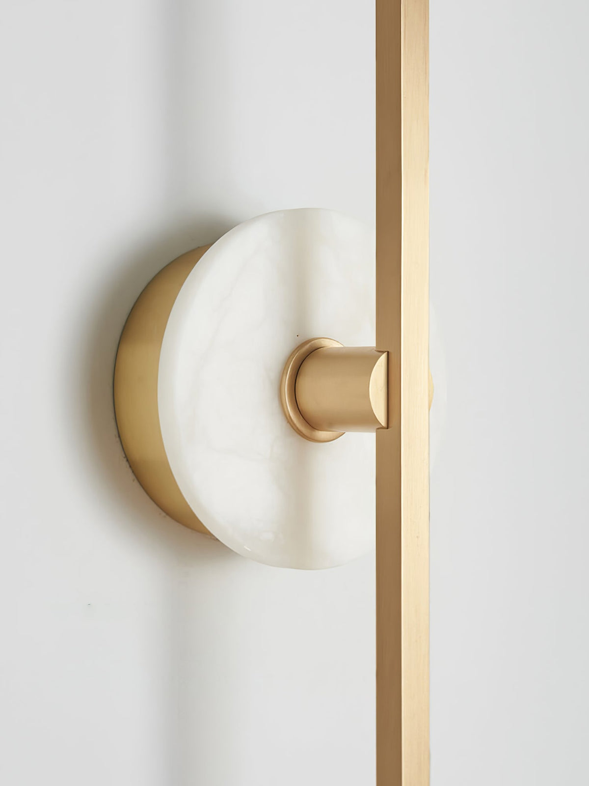 Close-up of a Clérachic wall sconce's pale alabaster disc where it meets the brass bar against a plain wall.