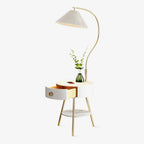 A Clérachic floor lamp arches its pleated white shade over a built-in round nightstand with a drawer, styled here with a small vase of greenery on top.