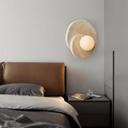 This Clérachic wall light glows above a leather-headboard bed, its stone discs catching soft light beside a cluttered nightstand.