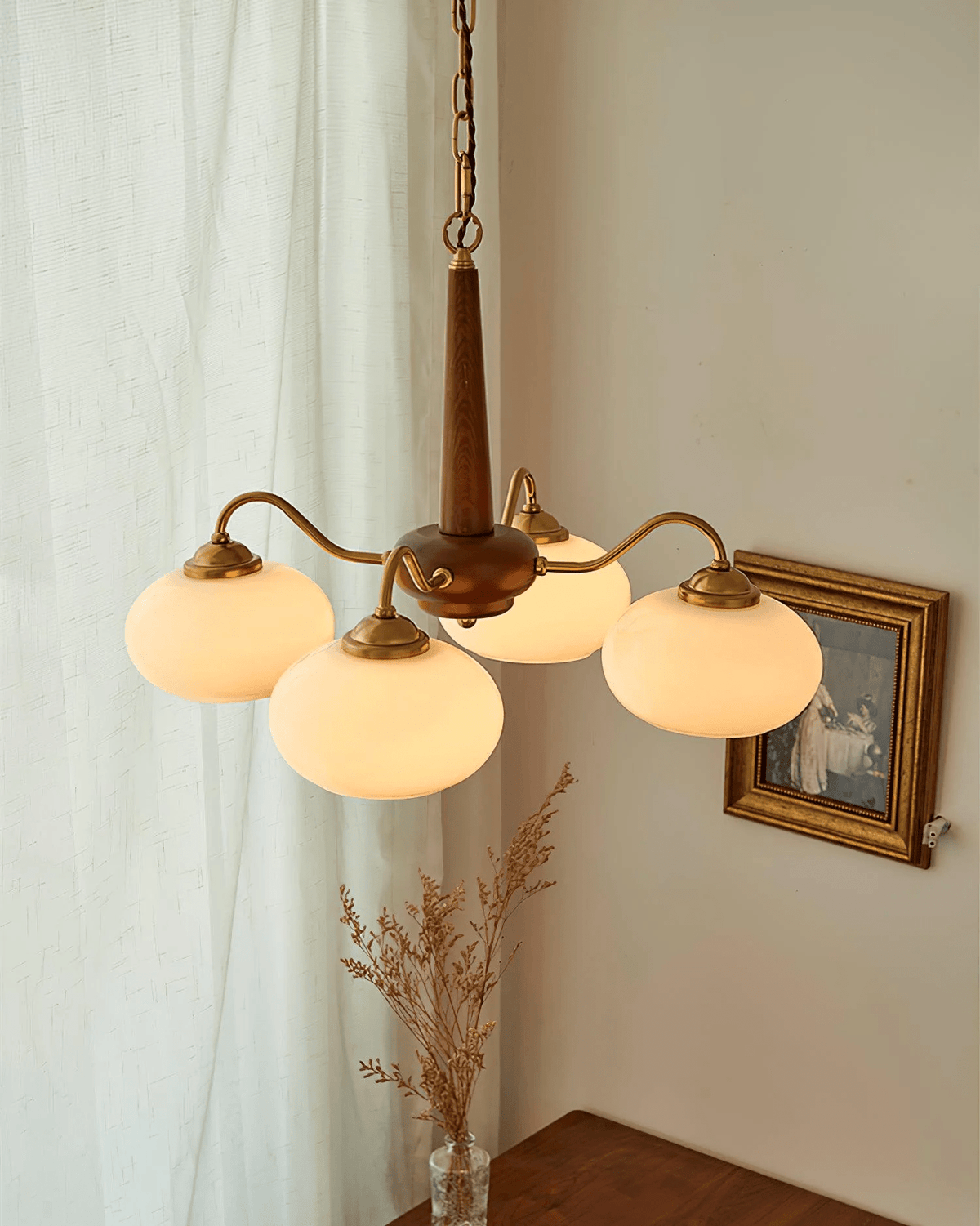 Dried grass in a glass vase sits beneath this Clérachic chandelier, its four cream shades lit near a curtain.