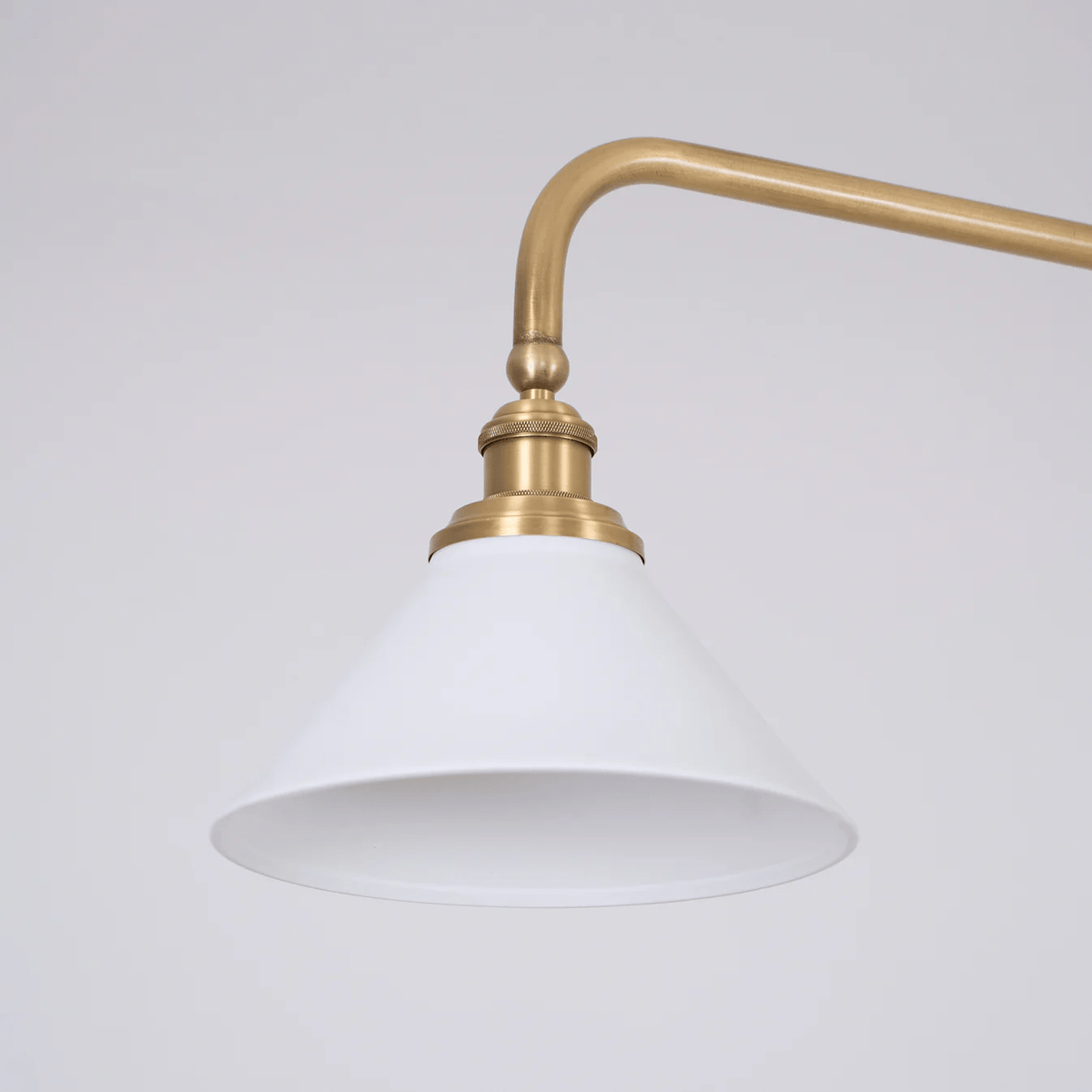 Close view of one white conical shade and its brass elbow arm on this Clérachic pendant, unlit against grey.