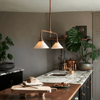 This Clérachic pendant glows warm above a marble kitchen island, its brass arms set against dark grey cabinetry and potted plants.