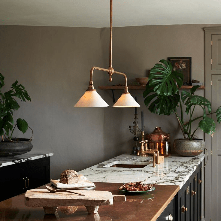 This Clérachic pendant glows warm above a marble kitchen island, its brass arms set against dark grey cabinetry and potted plants.