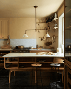 Wood open shelving and bar stools sit beneath this Clérachic pendant, glowing warm above a dark-floored kitchen counter.