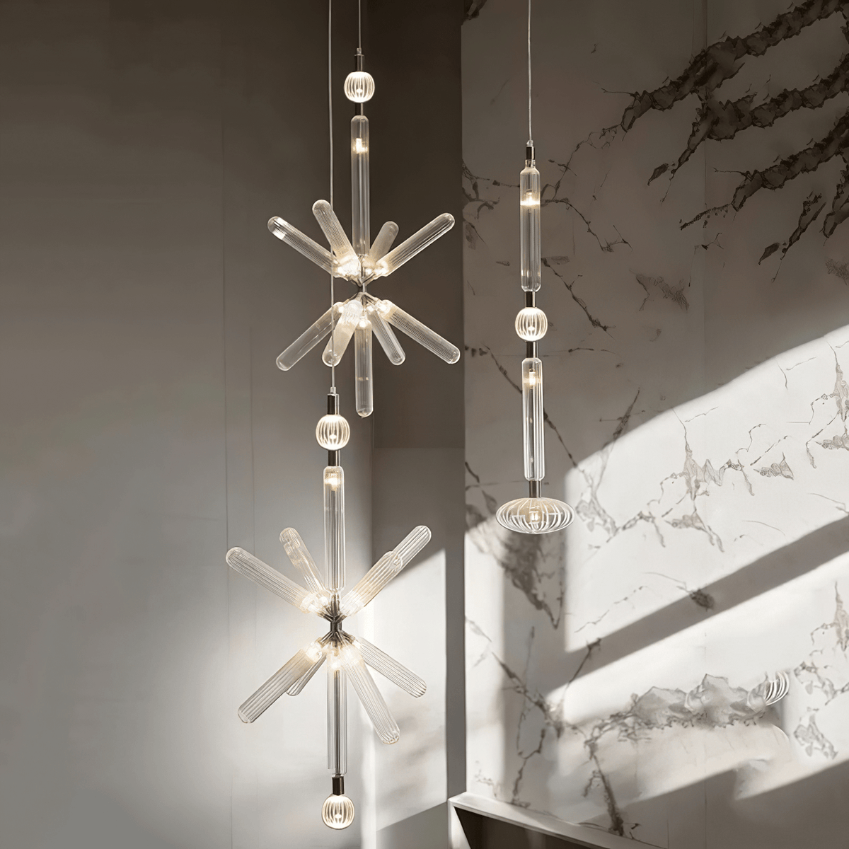 A crystal-burst Clérachic pendant and a linear one hang lit against a marble wall.