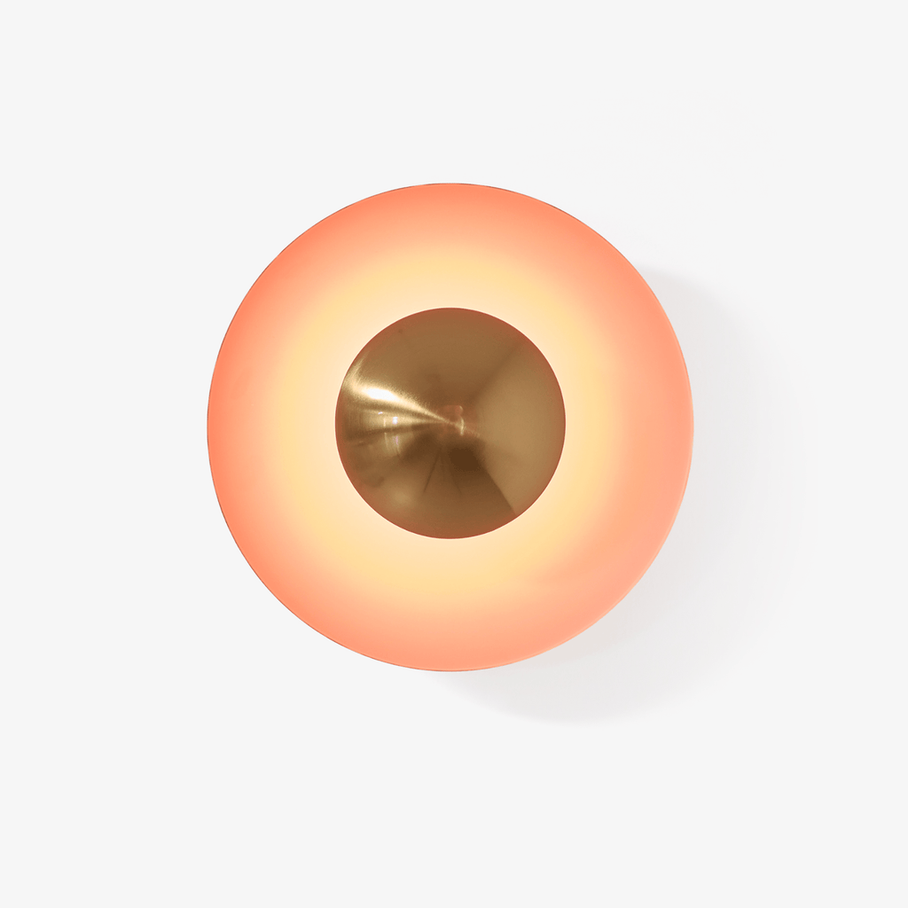 Close view of this Clérachic wall light in orange, its glowing shade set off by an antique copper centre, on a plain backdrop.