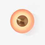 Close view of this Clérachic wall light in orange, its glowing shade set off by an antique copper centre, on a plain backdrop.