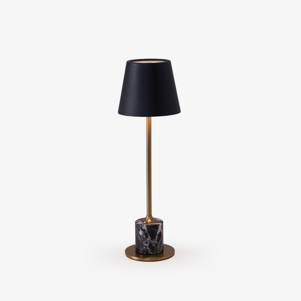 A slim brass stem rises from a black marble cylinder base to a black tapered shade on this Clérachic table lamp, photographed alone against a plain backdrop.