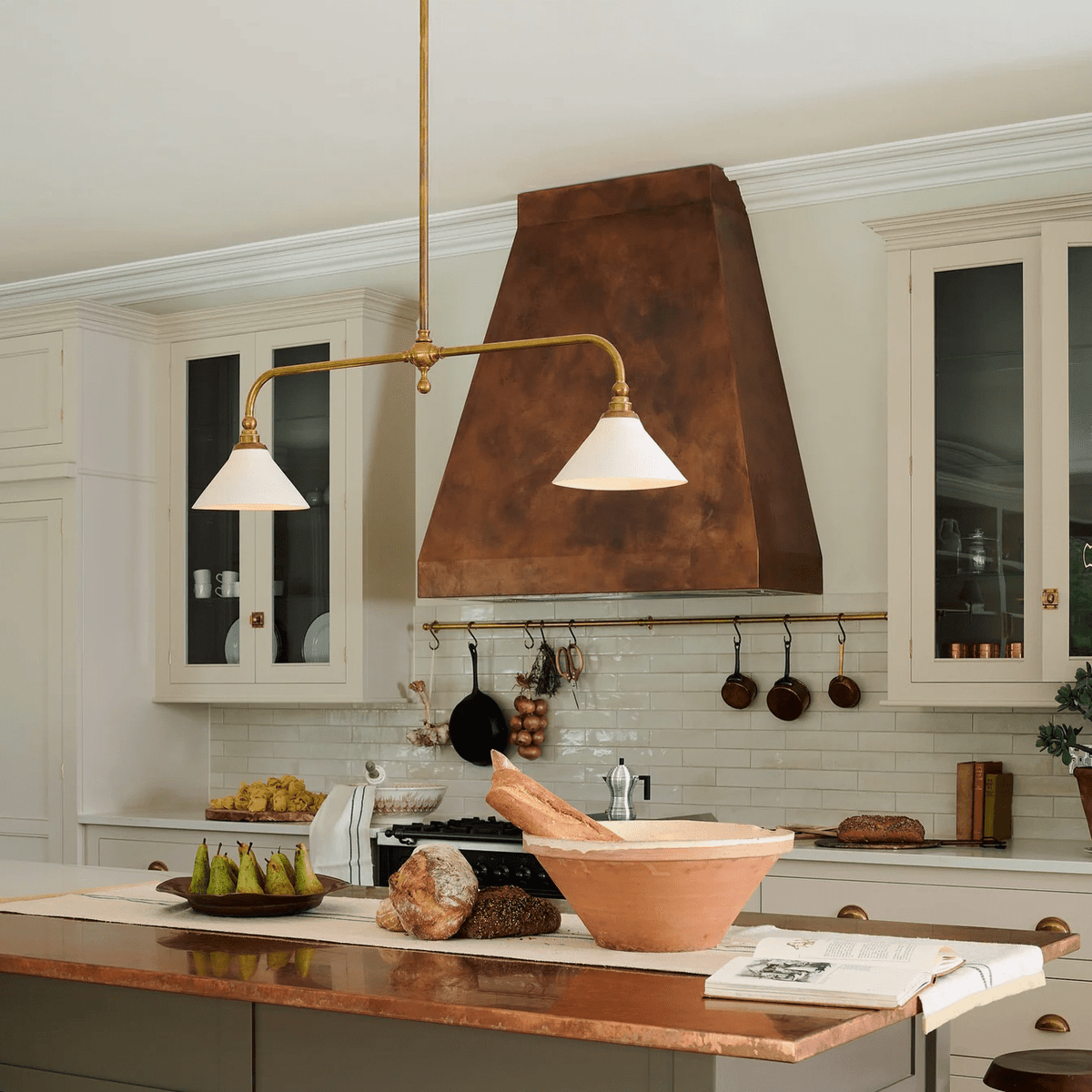 Beneath a copper range hood and white cabinetry, this Clérachic pendant glows warm above a kitchen island with fresh produce.