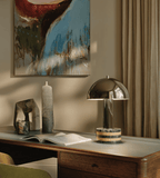 Clérachic table lamp on a wooden console beneath abstract wall art, small sculptural objects and an open book nearby.