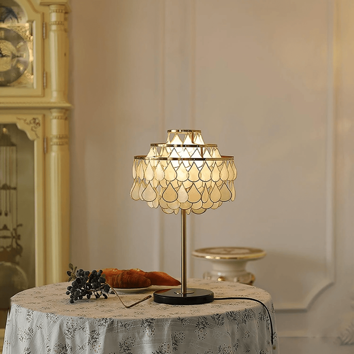 Clérachic table lamp glowing on a round floral-cloth table, a croissant plate and blueberries just beside its base.