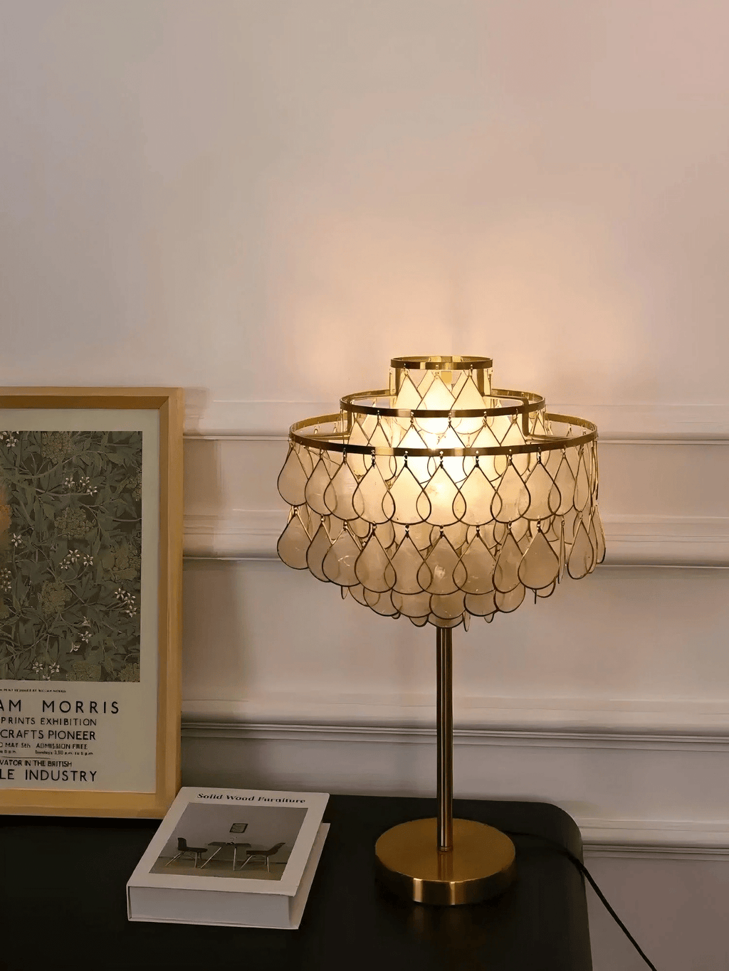 Near a gilt-framed picture on the wall, the Clérachic table lamp glows on a dark surface beside a small gift box.