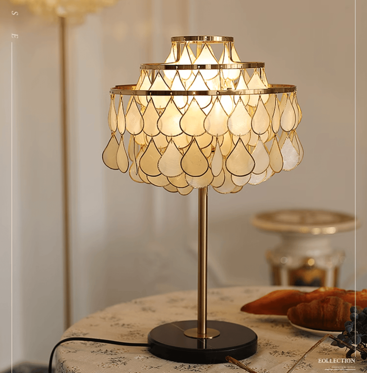 An ornate clock cabinet blurs behind the Clérachic table lamp, glowing on a round table with croissants and blueberries.