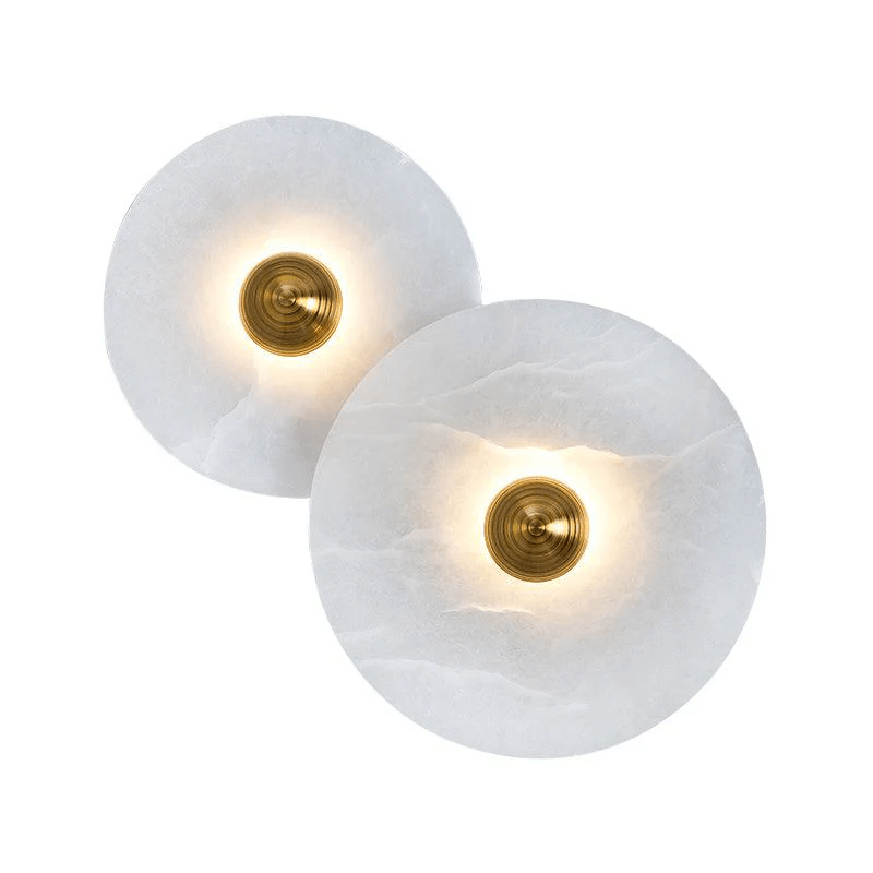 Pair of lit alabaster disc sconces with polished brass centres, glowing warm against white.