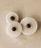 Three unlit alabaster discs on a warm beige plaster wall, dark bronze centres casting soft shadow.