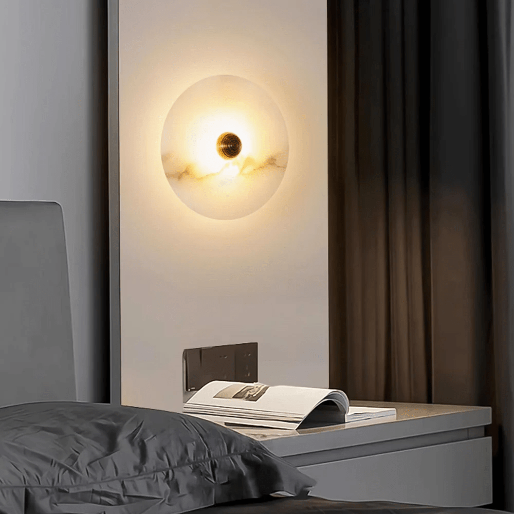 Alabaster disc sconce lit on a white wall panel above a bedside table with an open magazine.