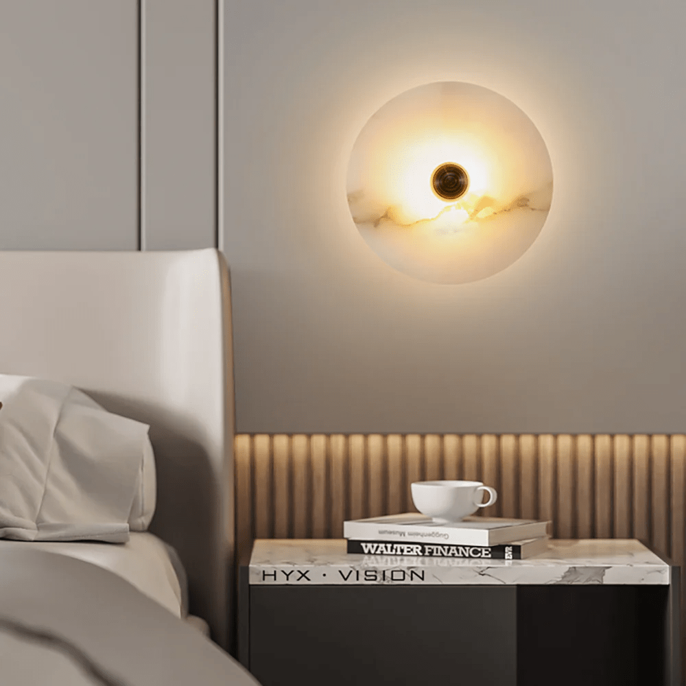 Round alabaster wall light glowing beside a bed, above a nightstand stacked with books and a cup.