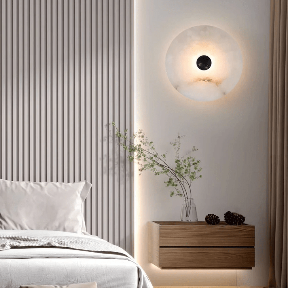 Alabaster disc sconce lit on a bedroom wall beside a wooden nightstand and a vase of branches.