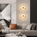 Two alabaster disc sconces stacked vertically and lit, on a living-room wall above a grey sofa.