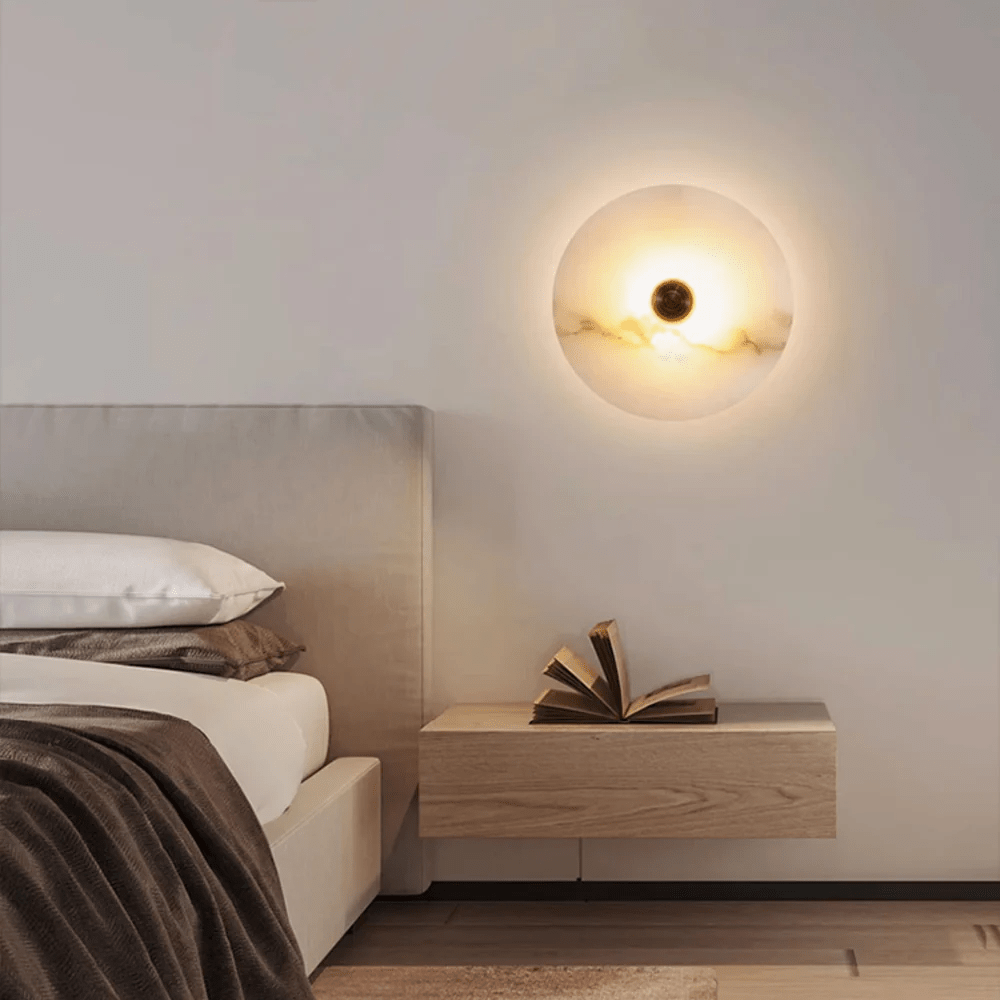 Round alabaster wall light lit above a wooden nightstand with an open book, beside a bed.