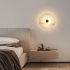 Round alabaster wall light lit above a wooden nightstand with an open book, beside a bed.