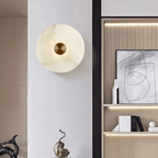 Unlit alabaster disc sconce on a white pillar wall, with shelving and artwork beyond.