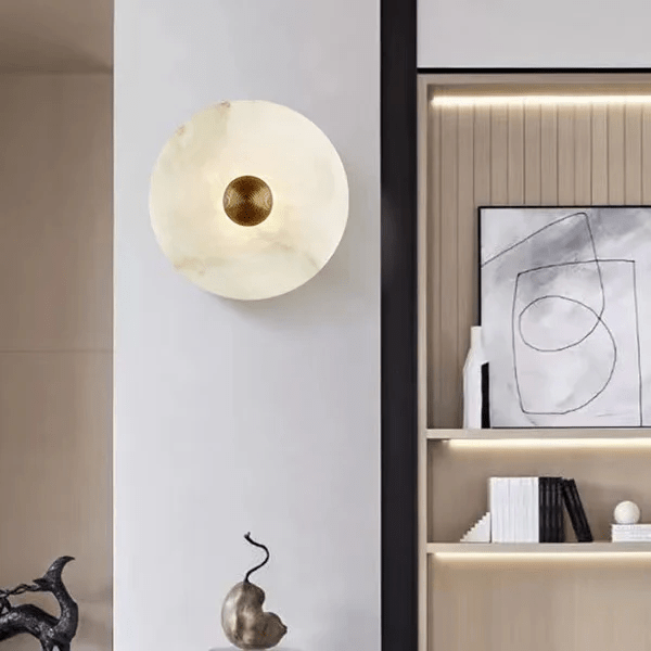Unlit alabaster disc sconce on a white pillar wall, with shelving and artwork beyond.