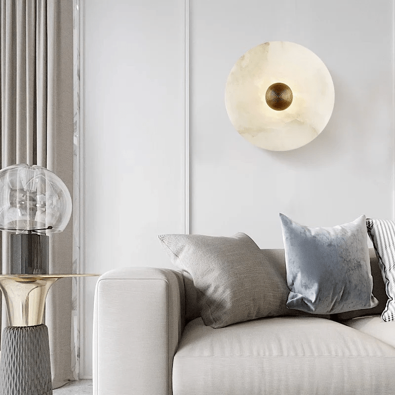 Alabaster disc wall light unlit on a panelled wall above a cream sofa with grey cushions.