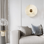 Alabaster disc wall light unlit on a panelled wall above a cream sofa with grey cushions.