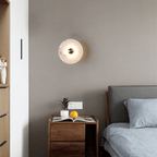 Alabaster disc sconce lit on a grey bedroom wall above a walnut nightstand.
