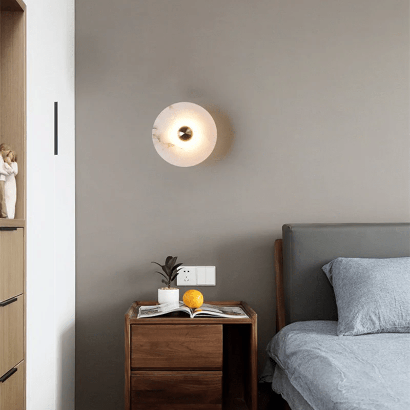 Alabaster disc sconce lit on a grey bedroom wall above a walnut nightstand.
