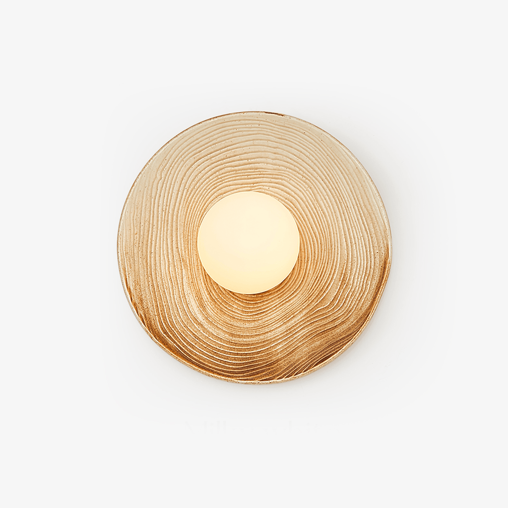 A ridged terra-beige ceramic wall sconce from Clérachic on a white studio background, its opal globe glowing warm off-centre.