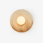A ridged terra-beige ceramic wall sconce from Clérachic on a white studio background, its opal globe glowing warm off-centre.