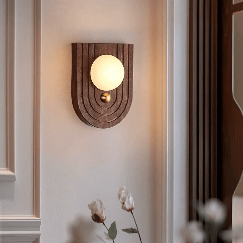 Arched walnut Koyan wall light lit on a panelled wall beside dried cotton stems.