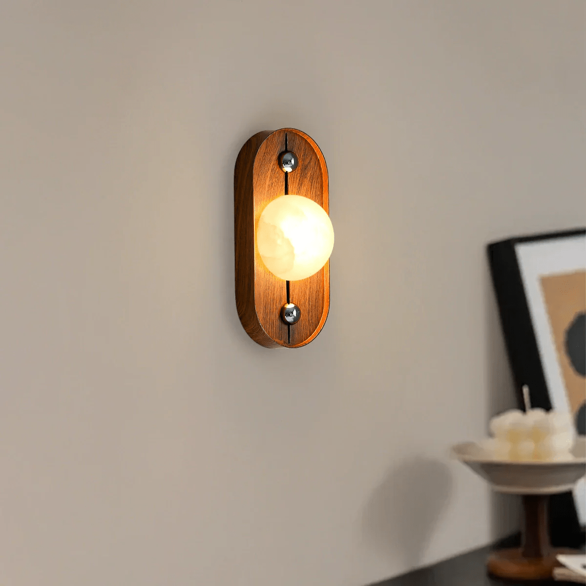 Oval Koyan wall light glowing on a taupe wall, seen at an angle beside framed art.