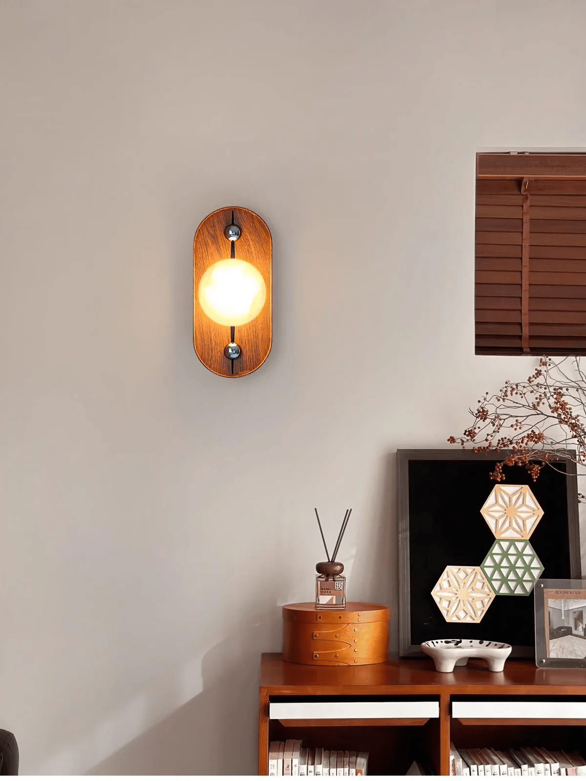 Oval Koyan wall light lit above a walnut sideboard, wooden blind and hexagon artwork nearby.