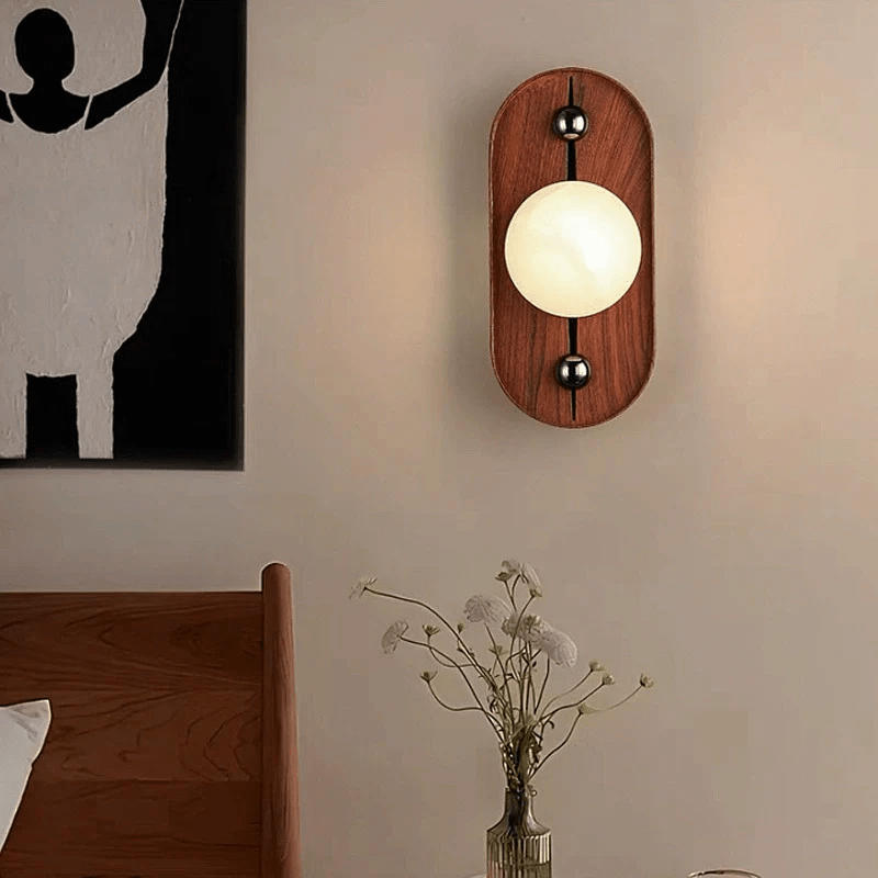 Oval Koyan wall light in a bedroom, mounted above a wooden bed beside a monochrome print.