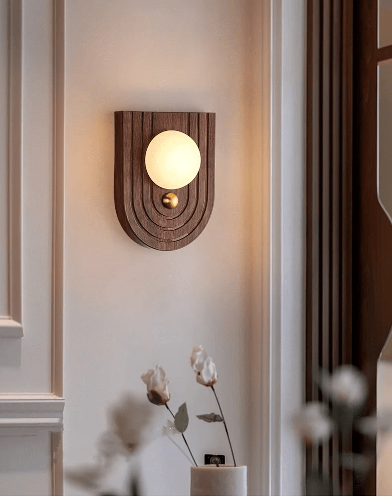 Arched walnut Koyan wall light warming a moulded panel wall, dried cotton blooms in the foreground.