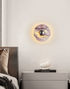 Clérachic's wall light with a gunmetal centre knob glows above a leather headboard, beside a grey nightstand and small figurine.