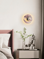 A lit Clérachic wall light glows above a mauve upholstered headboard, beside a glass vase of eucalyptus stems and a figurine.