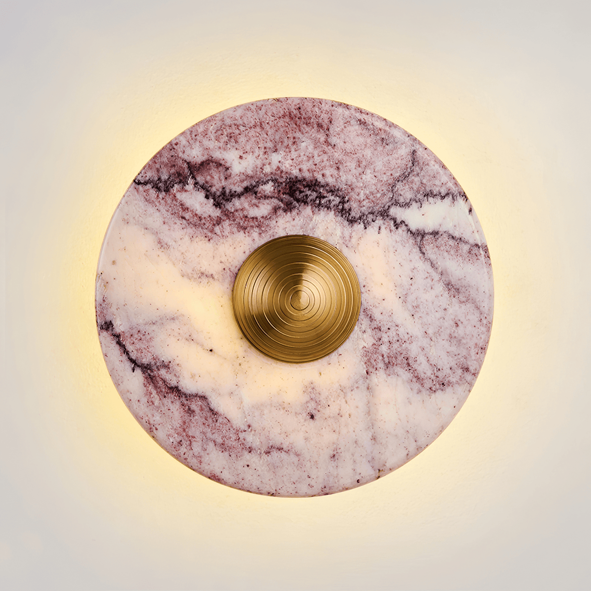 Close-up of a Clérachic wall light in purple-veined marble, its gold ridged centre knob glowing warm against a soft backlit halo.