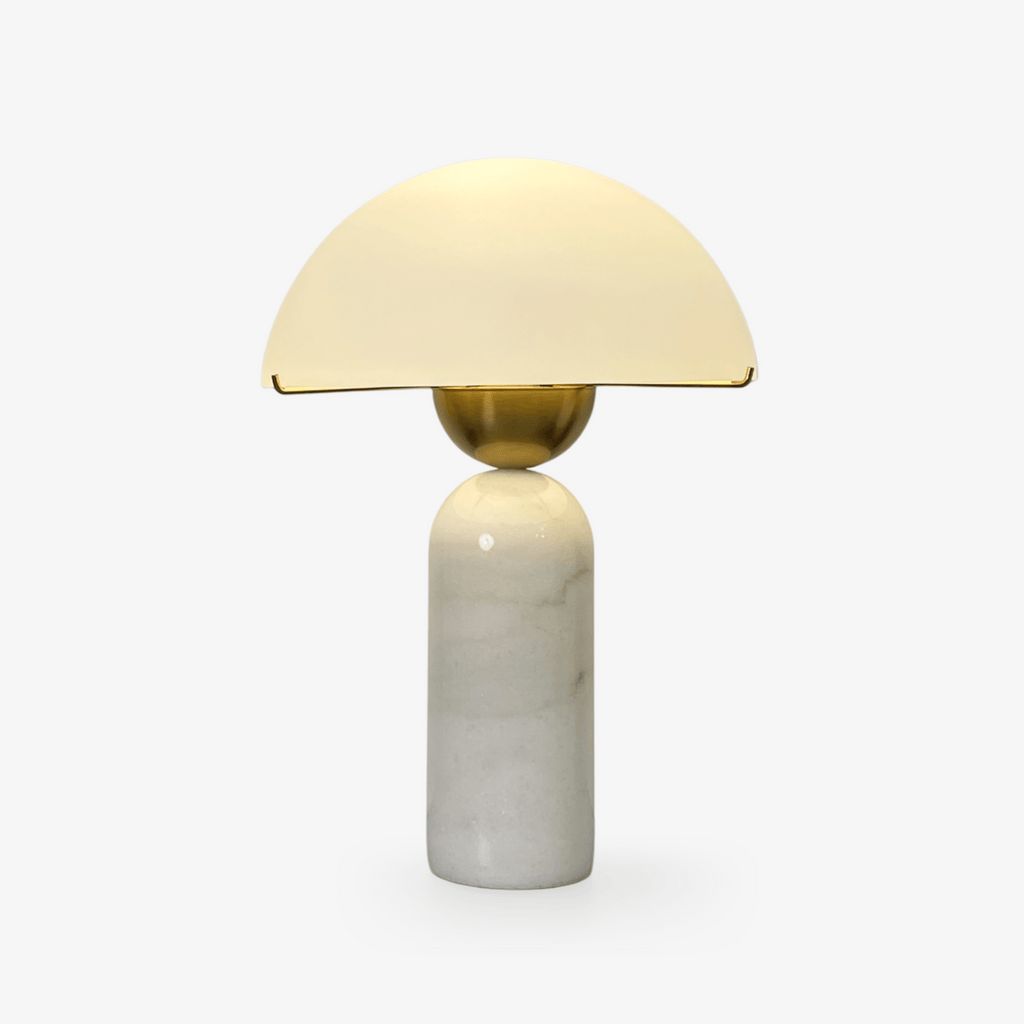 A Clérachic dome table lamp with a white marble base and gold sphere stem glows softly on a plain light backdrop.