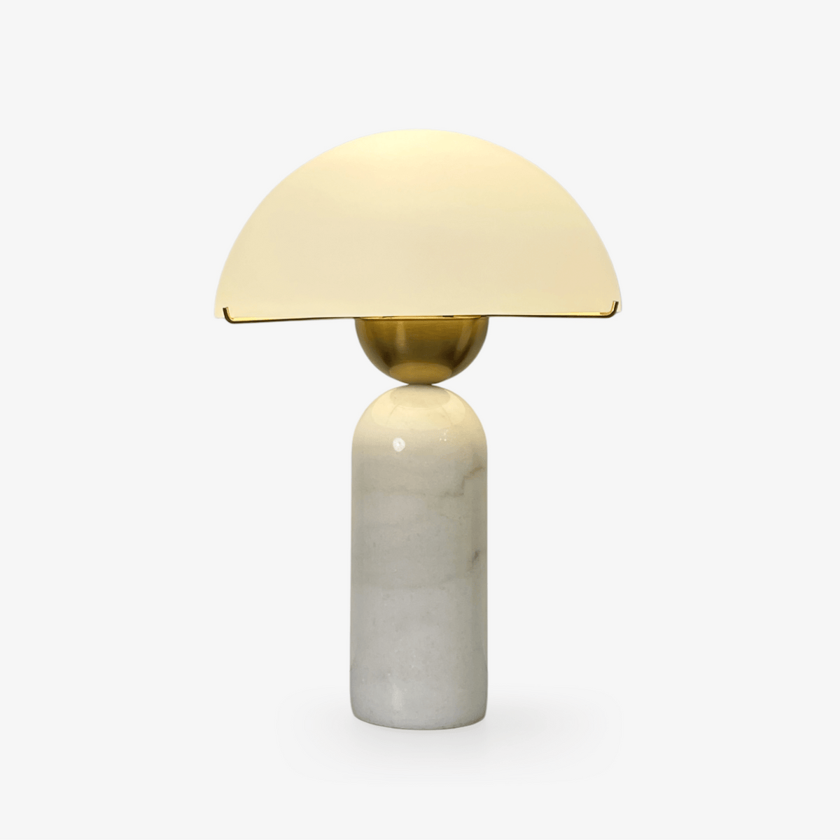 A Clérachic dome table lamp with a white marble base and gold sphere stem glows softly on a plain light backdrop.