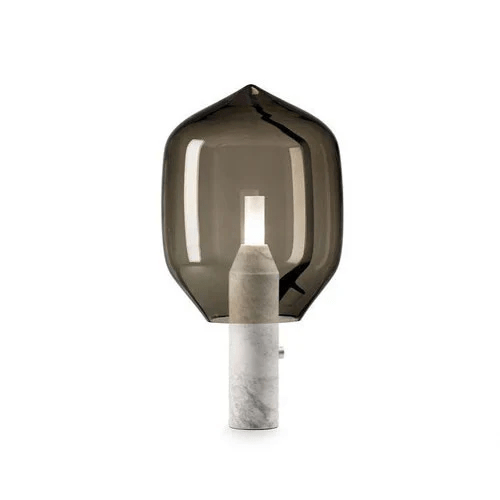 A smoked-glass Clérachic table lamp on a white marble base, lit against a plain grey-white studio backdrop.
