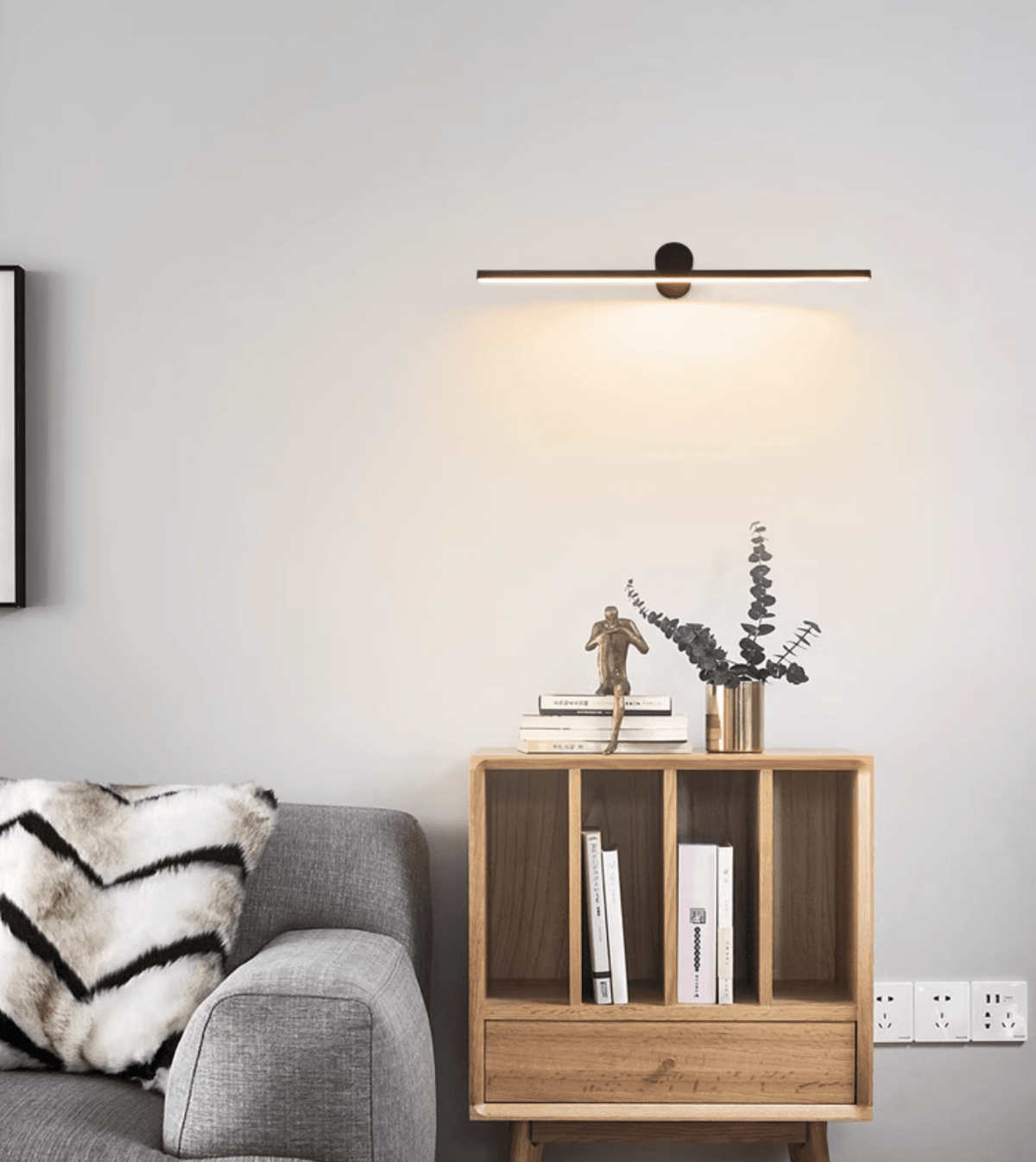 This Clérachic wall light glows above a wood bookshelf holding dried greenery and stacked books.