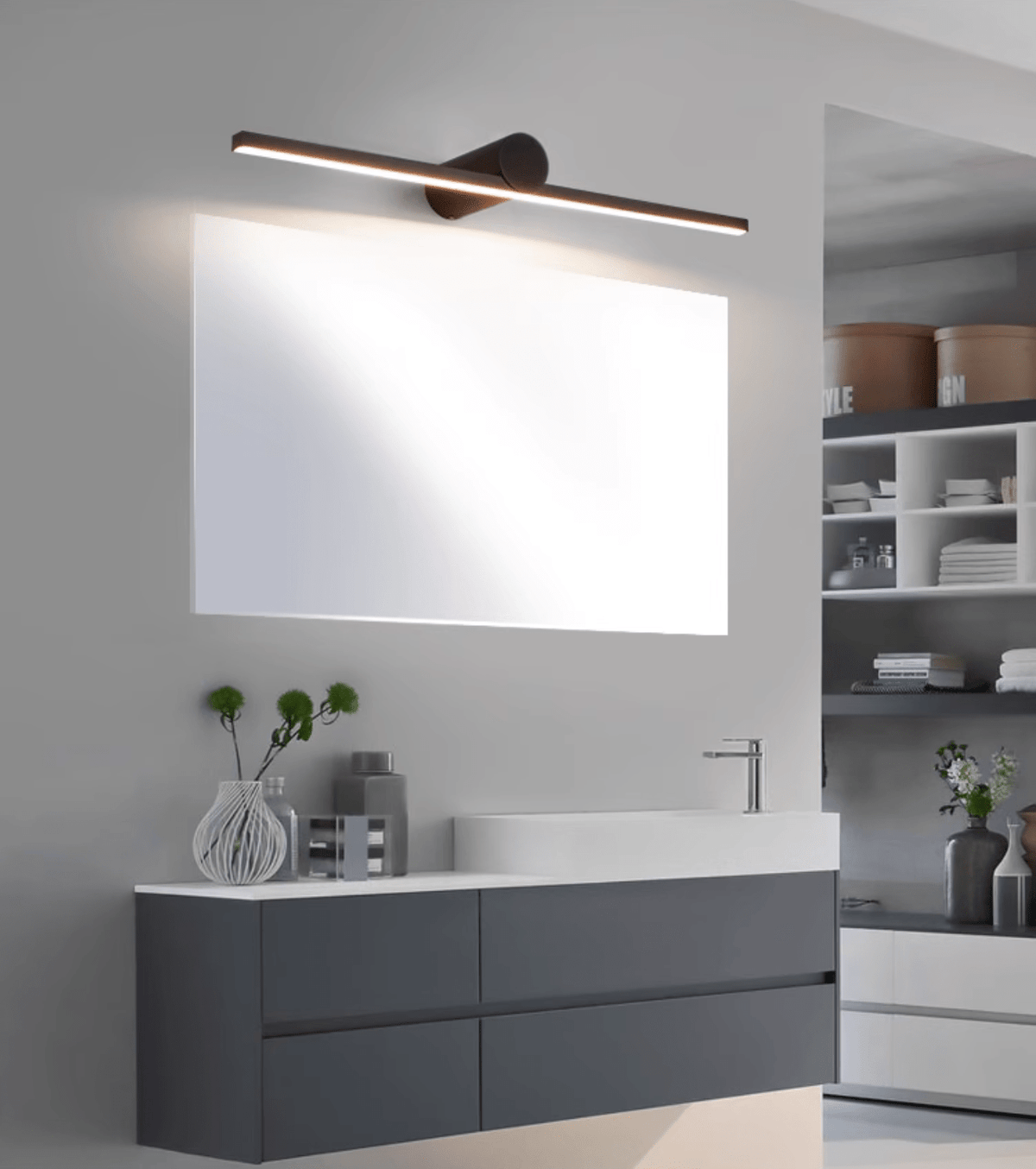 A lit Clérachic wall light spans a wide mirror above a grey bathroom vanity with a vase of greenery.